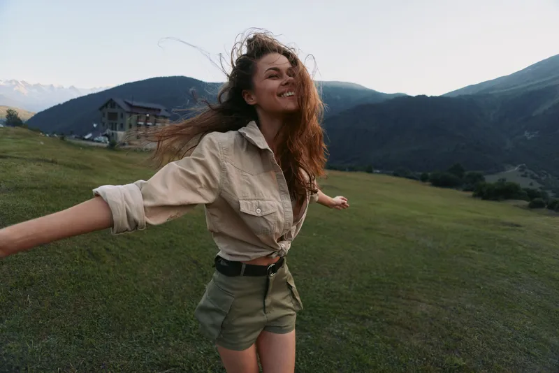 Smiling woman with long hair wearing a beige shirt and khaki shorts in a grassy field, capturing an outdoor lifestyle moment filled with happiness and nature. Bright mood and color