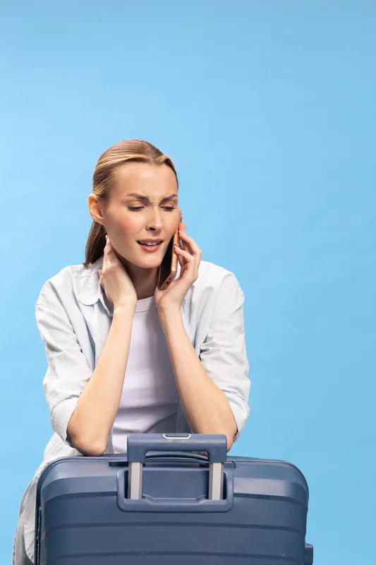 Young woman with suitcase talks on phone looking worried, isolated on blue background. Concept of travel problems, stress, and delayed flights or cancellations.