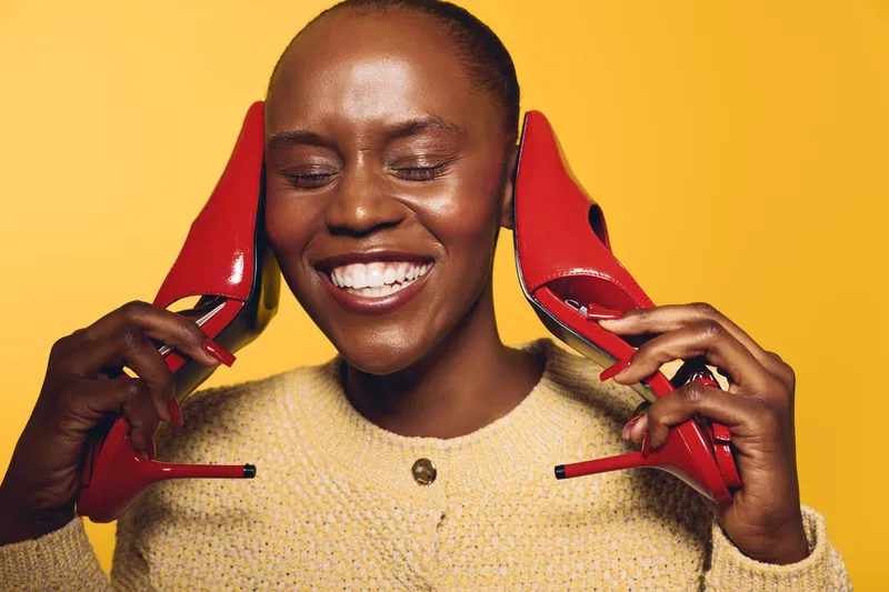 Smiling black woman with dark skin holds red high heels near her face against yellow background. Concept of fashion, joy, beauty, and confident female model with stylish shoes.