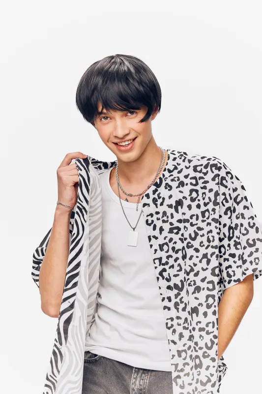 Young man smiling confidently in trendy patterned shirt holding jacket over shoulder, styled black hair, casual white t-shirt, isolated white background.