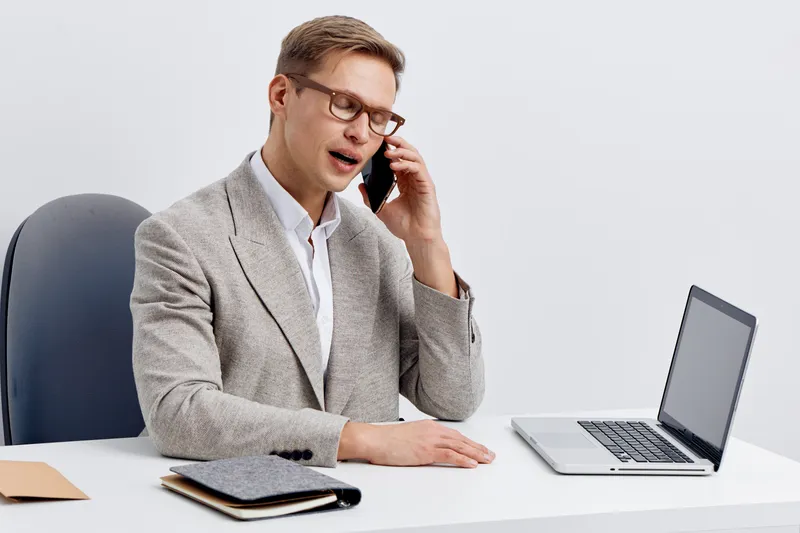 Professional man in casual office attire talking on mobile phone while sitting at desk with laptop, notebook, and documents. Confident, focused expression. Studio shot on plain white background. Busin