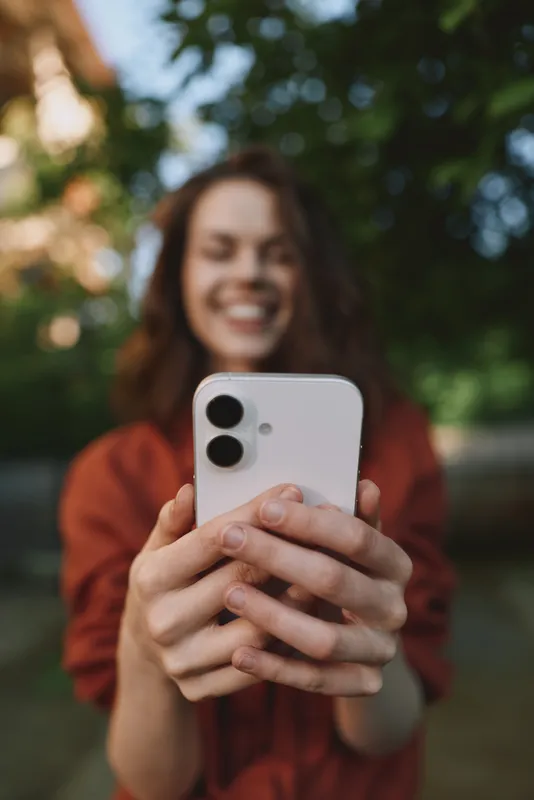 Joyful woman outdoors, smiling while taking a selfie, warm sunlight, casual outfit, blurred background of trees, candid lifestyle moment, natural colors, relaxed posture, fresh air, vibrant atmosphere