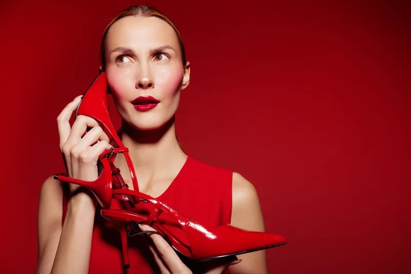 Stylish woman in red dress holds red high heel shoe near her face against red background. Concept for fashion, style, glamour, and bold color contrast in studio lighting.