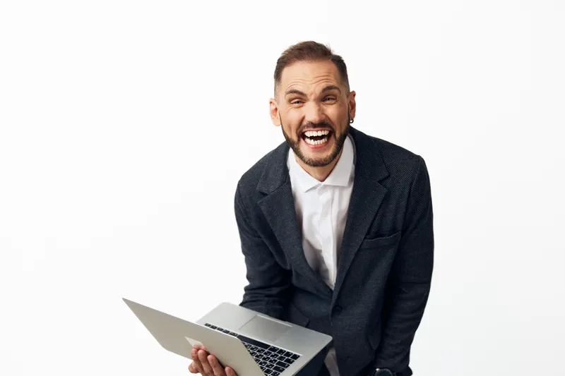 Professional business man with a laptop stands against a clean colored background, projecting energy, confidence, and excitement through his expressive smile.