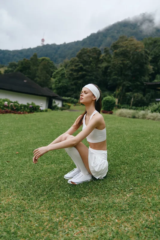 Young Asian woman in sporty white outfit and headband squatting on green grass, enjoying outdoor exercise in a lush natural environment