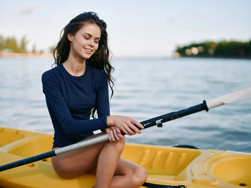 A young woman sits in a yellow kayak by calm water, wearing a navy long sleeve top and shorts, holding a paddle with a gentle smile.