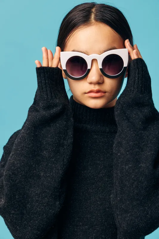 Young woman wearing oversized white sunglasses and black sweater posing against blue background with serious expression and hands near head, stylish fashion portrait.