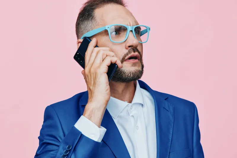 A professional man in a blue suit speaks on a mobile phone against a solid pink background, conveying focus, confidence, and modern business style in a clean studio setting.