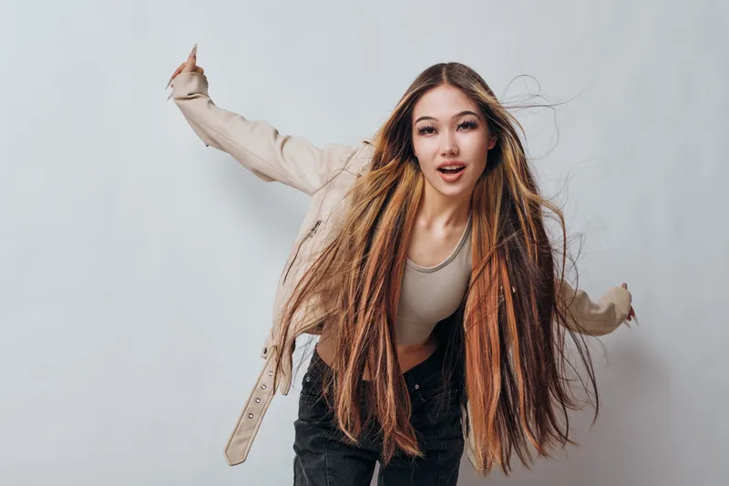 Young woman with long hair wearing beige jacket and light top posing with dynamic movement against a plain light background in studio. Casual style and confident expression.