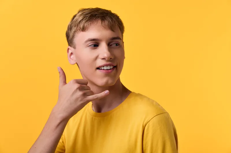Young man in a yellow tee making a phone gesture for communication against a bright studio backdrop, casual style, friendly mood, modern lifestyle and positivity
