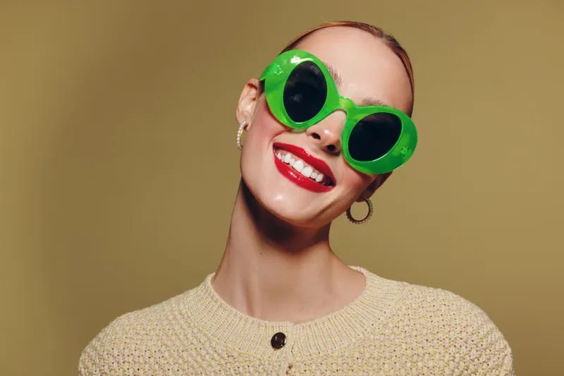 Smiling woman with bright red lipstick wears large green sunglasses and beige sweater. Studio portrait on neutral background. Concept of fashion, style, and confident female model.