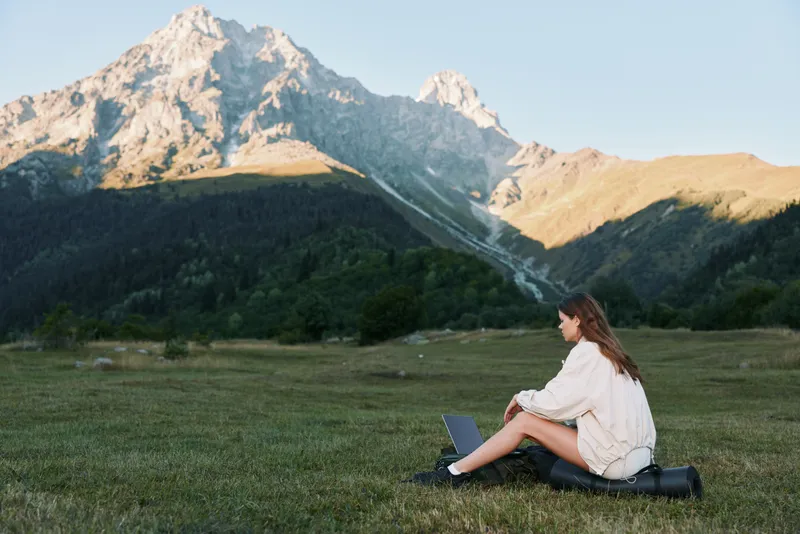 A young woman sits on a grassy meadow with a laptop in a remote mountain landscape, focused and calm as she works, enjoying solitude and a serene, productive atmosphere.