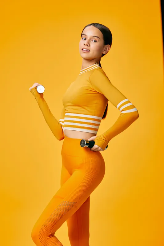 Woman fitness enthusiast in vibrant orange athletic wear holds a dumbbell, mid workout, against a bright orange backdrop, displaying training, strength and sport confidence.