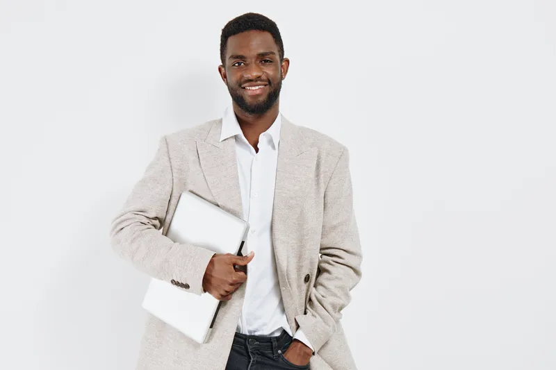 Confident smiling man holding laptop and notebook, dressed in beige blazer and white shirt, standing against plain white background. Studio portrait of professional businessman, casual style, positive