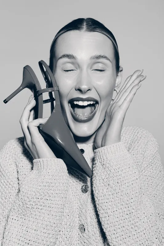 Happy white woman with light skin laughs joyfully holding high heel shoes near face in cozy sweater. Concept of fashion, happiness, and casual style in studio portrait.