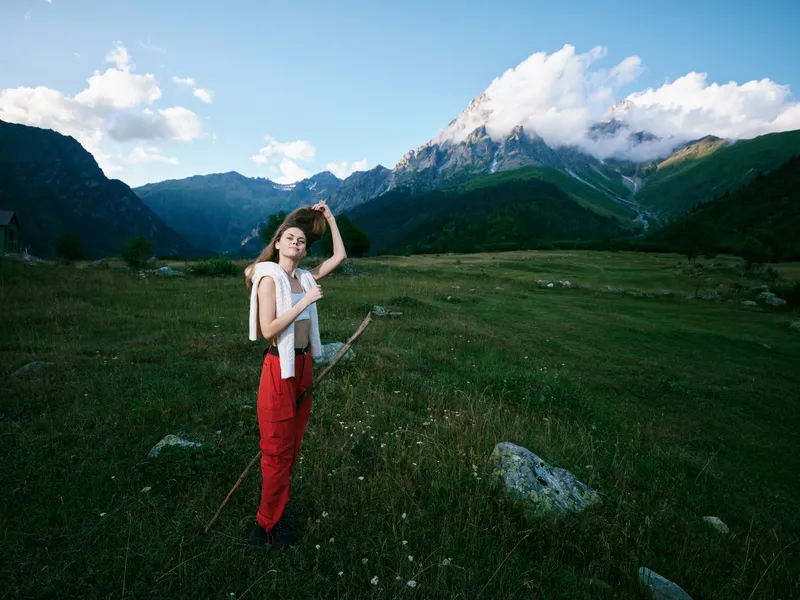 Hiker standing in a sunny meadow with distant mountains, backpack and walking stick, exploring remote terrain and enjoying expansive alpine scenery under a bright blue sky