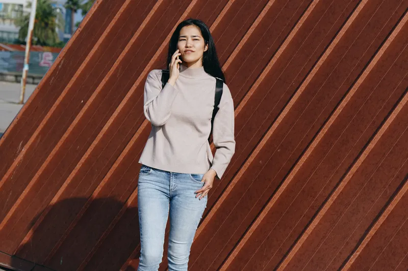 Phone in hand, casual outdoor scene with a smiling woman in jeans and sweater against a diagonal wooden wall, urban lifestyle and street fashion vibe