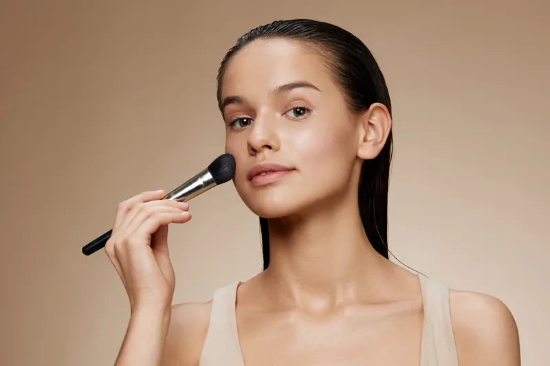 Young woman applying makeup with a brush, showcasing a natural beauty look against a neutral background, perfect for beauty and cosmetics themes