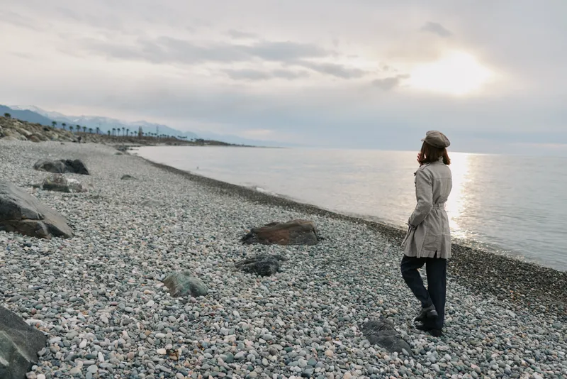 Woman in coat and hat walks alone on rocky beach during cloudy sunset. Calm sea and distant mountains create peaceful and contemplative mood in natural outdoor landscape.