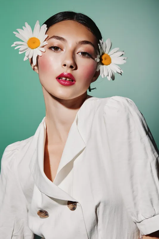 Fashion model wearing a white blazer with buttons and large white flowers in her hair posed against mint green background. Creative beauty concept with shiny red lips and radiant skin.