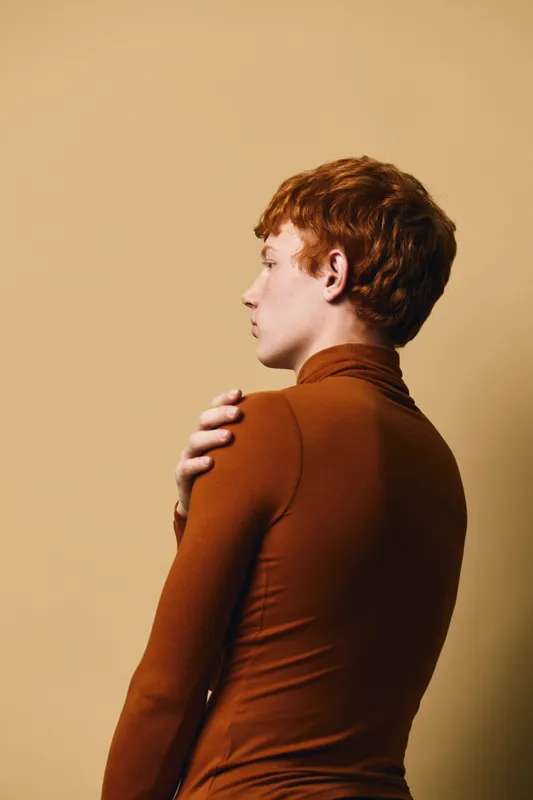 Redhead woman in brown turtleneck posing sideways with hand on shoulder on beige background for fashion, portrait and beauty concept with thoughtful expression and soft lighting.