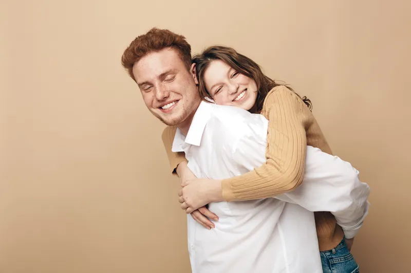 Happy young couple with closed eyes smiling while hugging each other on beige background. Concept of love, friendship, relationship, joy, and casual lifestyle together.