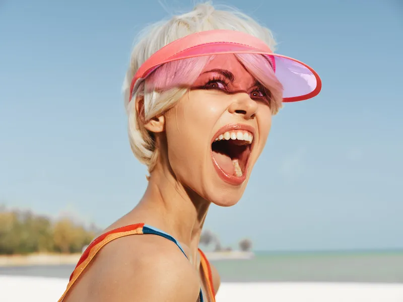 Vibrant beach scene featuring a young woman with pink hair and a pink visor, shouting with joy as the blue sea and sandy shore frame a dynamic summer moment.