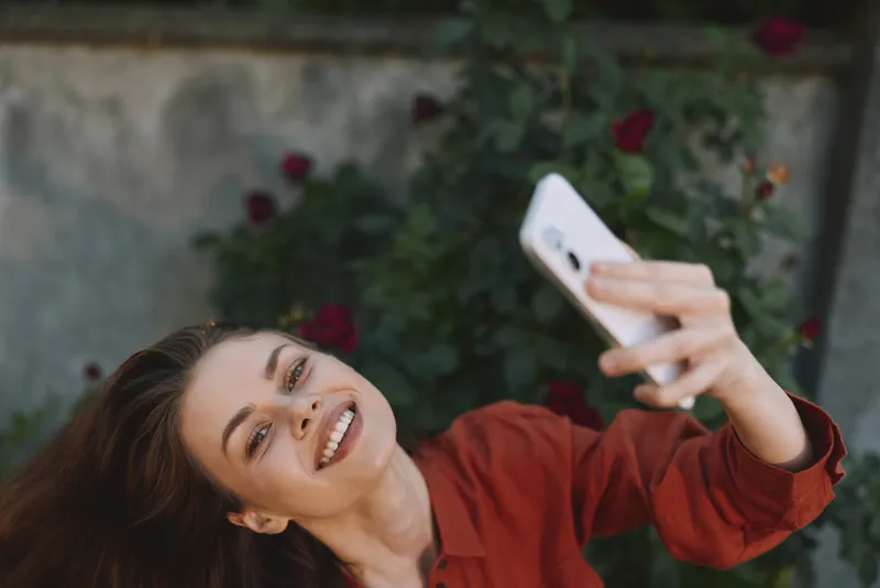 Bright woman takes a cheerful selfie outdoors in a garden filled with roses, wearing a red shirt, capturing a candid lifestyle moment and a warm natural smile for an authentic everyday scene