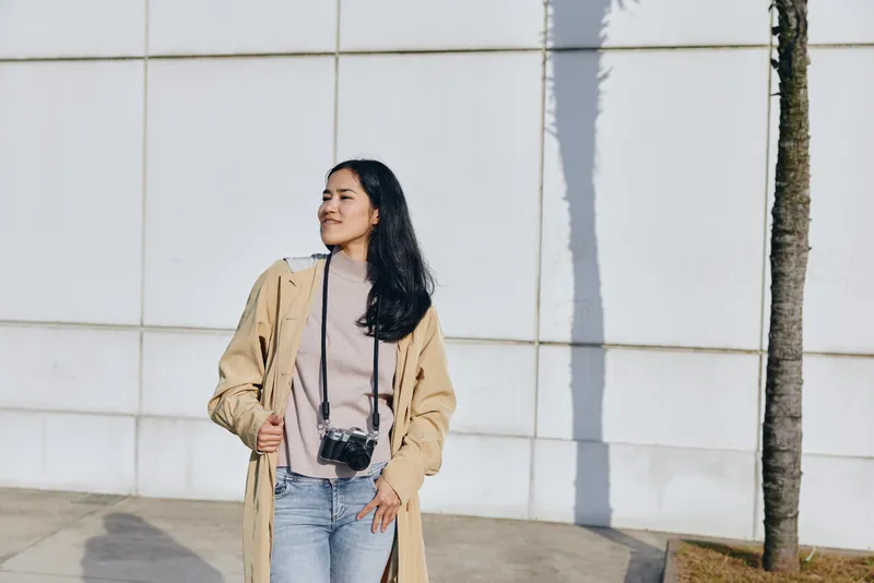 Woman wearing a beige trench coat and jeans with a camera hanging from her neck stands on a sunny street beside a wall, showcasing casual fashion and street photography vibe