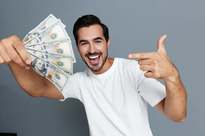 Smiling man in white t-shirt holding cash, pointing with finger at camera in studio against gray background. Concept of success and happiness.