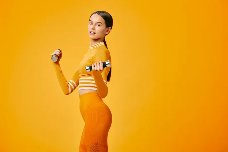 Fitness focused woman in a bright orange studio outfit, holding dumbbells and performing a dynamic workout with energy, balance and strength. The athletic pose captures mid-motion, showcasing a lean