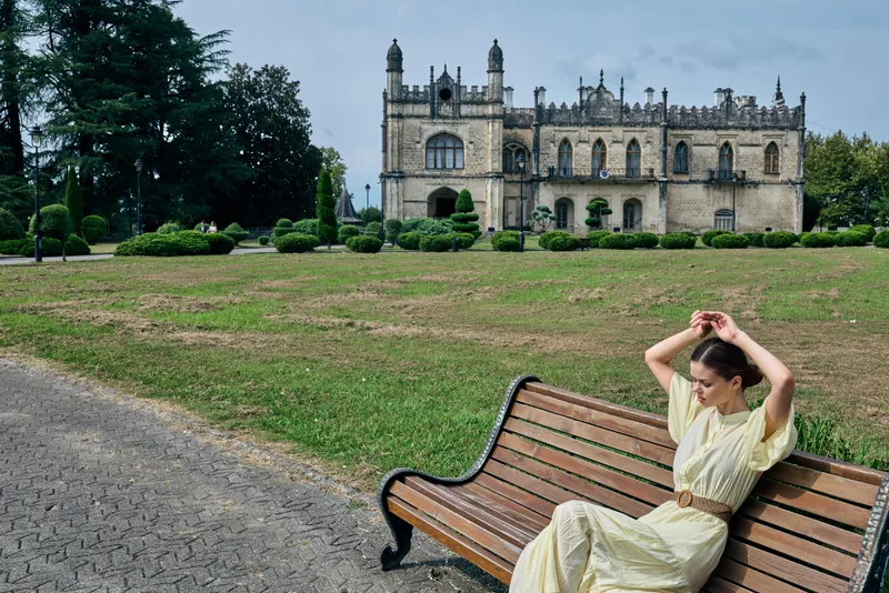 Woman in a light dress sits on a wooden bench in a park with a grand historic castle in the background, creating a serene travel and sightseeing moment.