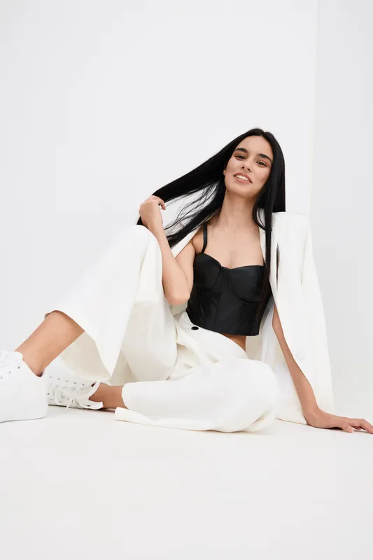 smiling woman with long black hair wearing white pants and jacket with black top sitting relaxed against white background, casual fashion look