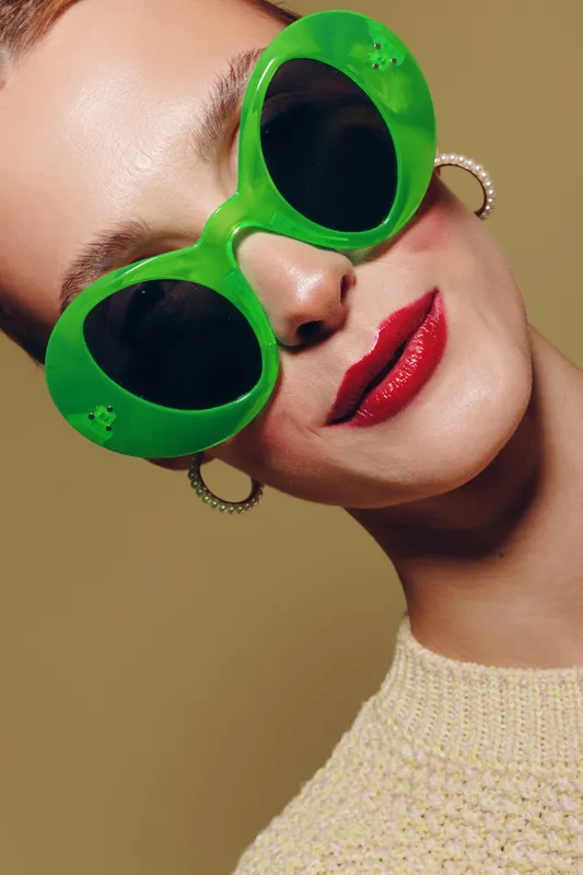 Close-up of a woman wearing oversized green sunglasses and red lipstick. She has hoop earrings and a beige knitted top. Concept of fashion accessories, beauty, and summer style.