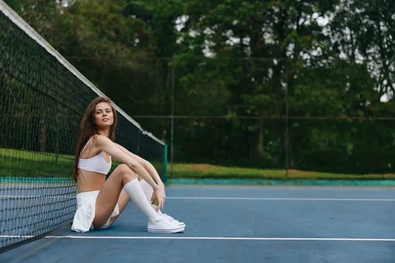 Young female tennis player sitting on the court, wearing a sporty outfit, smiling, enjoying a sunny day outdoors, with green trees in the background