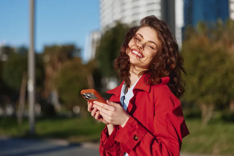 A happy young woman in a red coat smiles while holding a smartphone outdoors on a sunny day with blurred city buildings in the background.