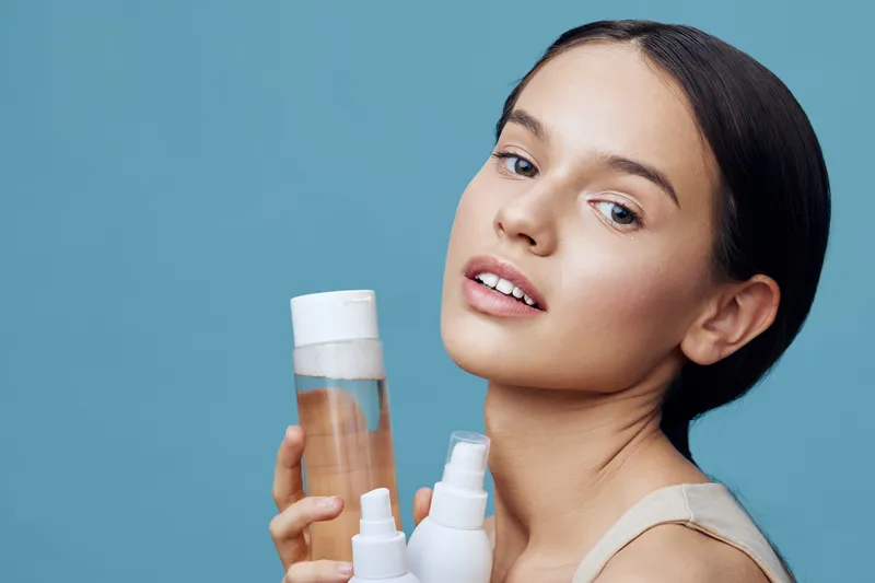 Young woman showcasing skincare products against a blue background, exuding confidence and elegance with a fresh and natural look