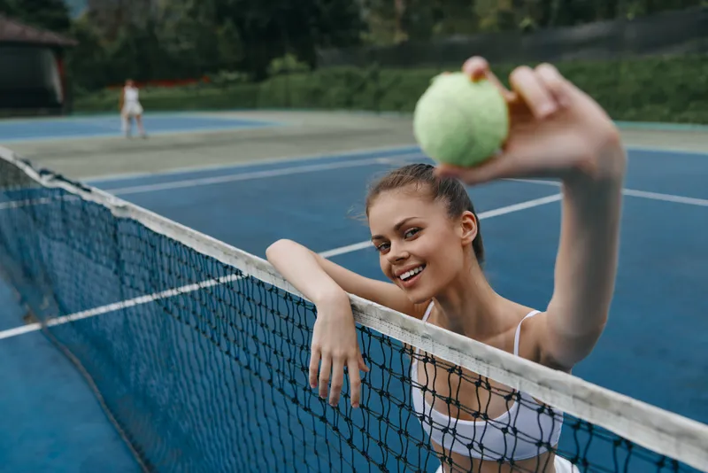 Young female tennis player smiling and holding a tennis ball at a blue outdoor court, showcasing athletic wear and positive energy during summer practice