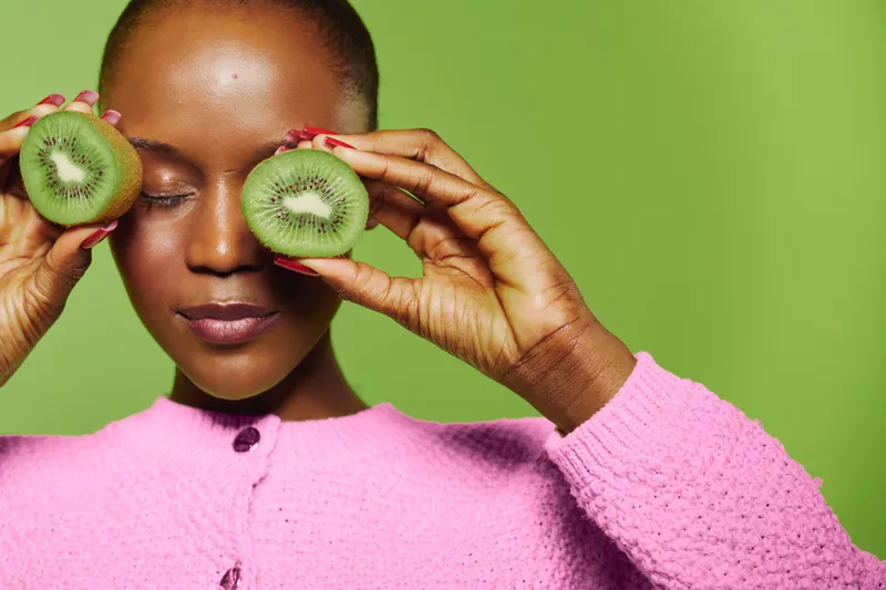 Dark skinned African woman with closed eyes holds two halves of kiwi fruit near her face, wearing pink sweater. Concept of natural beauty, healthy lifestyle, and fresh fruit skincare.