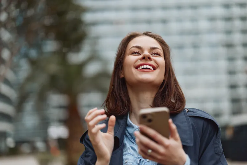 Young woman with long brown hair smiles happily while holding smartphone outdoors in casual jacket, enjoying sunny day in urban environment with blurred background.