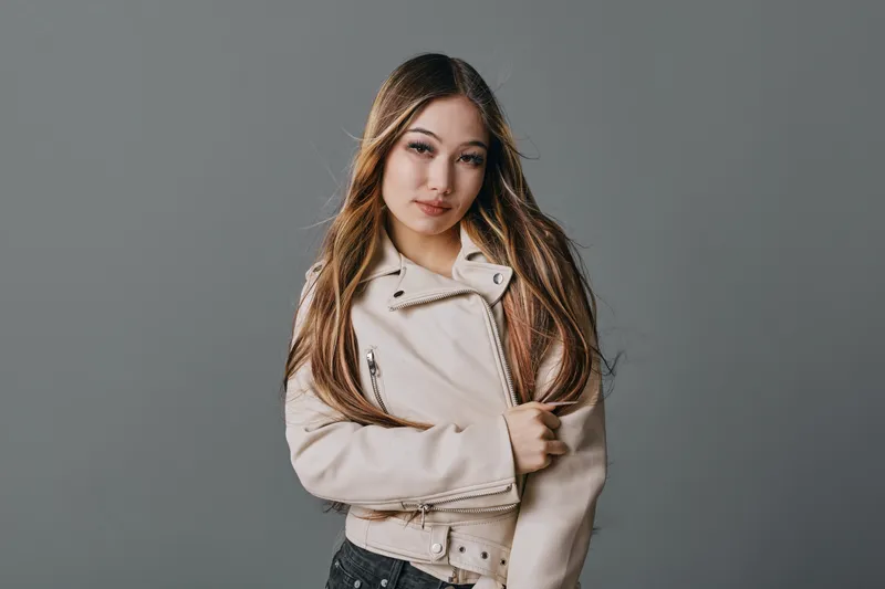 Young woman with long hair wearing light jacket posing against gray background looking at camera with confident expression in studio portrait with casual style and natural makeup.