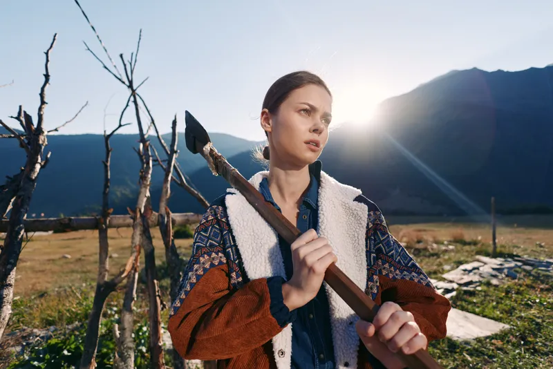 Woman with axe outdoors in a rural field by mountains at sunrise, portrait of a determined lumberjack holding tool over shoulder, natural light and rustic landscape.