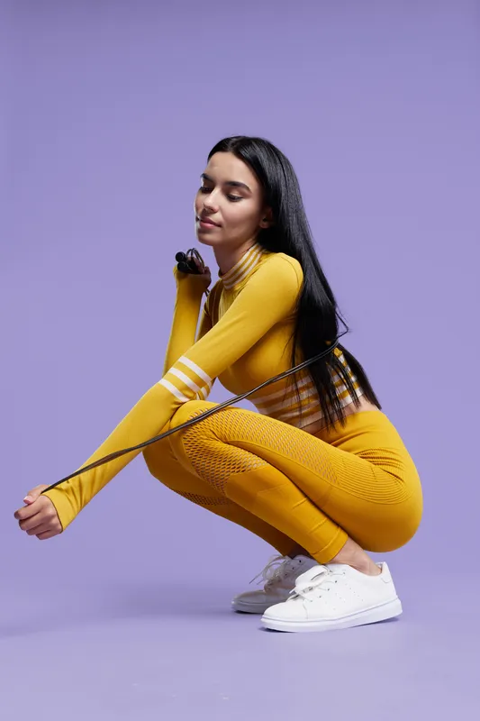 woman sport fitness dressed in yellow activewear squatting on purple background, holding a jump rope, focused and healthy lifestyle concept