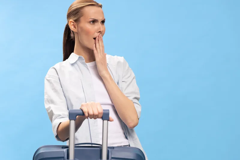 Worried woman with suitcase covers mouth with hand, looking aside on blue background. Concept of travel anxiety, uncertainty, and last minute problems in airport or trip planning.
