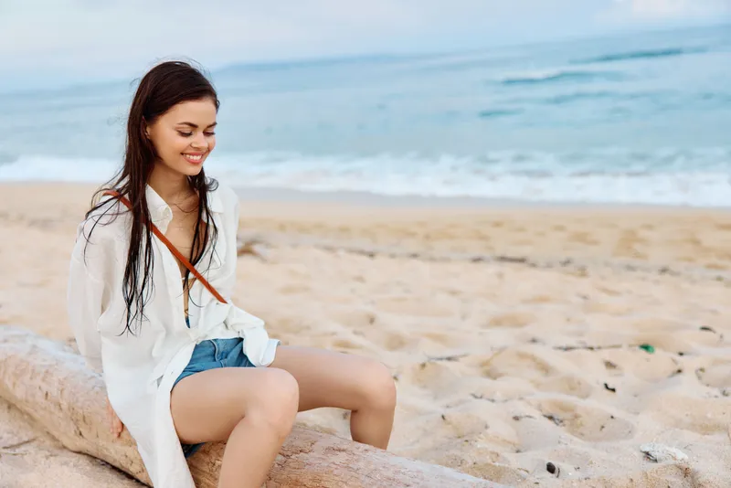 Счастливая загорелая женщина, улыбающаяся с зубами в белой рубашке-купальнике и джинсовых шортах, сидит на песке у океана с мокрыми волосами после купания, свет заката и розовые облака на Бали