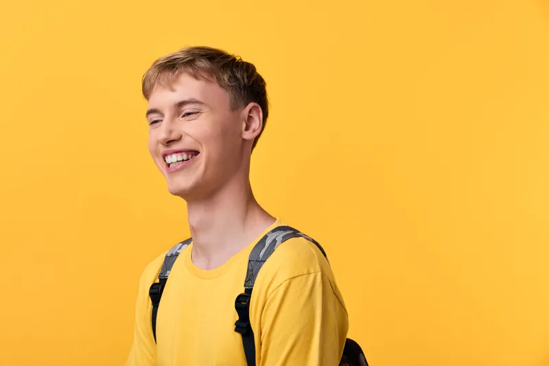 Smiling man with a backpack and yellow shirt against a bright background, displaying casual fashion, friendly vibe, and positive mood for lifestyle, education, youth, travel and daily activities