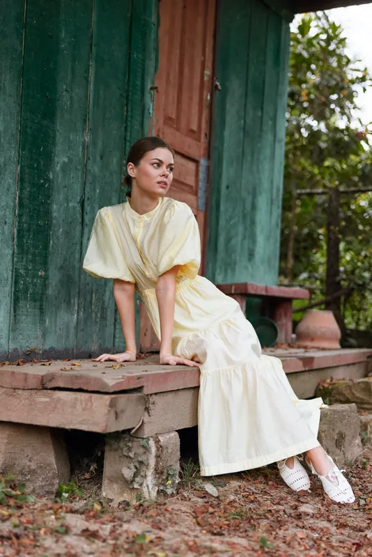 Woman wearing a pale cream dress sits on weathered wooden steps beside a green wall, posing gracefully outdoors, exuding calm elegance and refined fashion in a rustic setting.