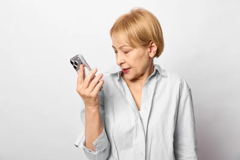 Elderly woman, smartphone, concerned expression, screen gaze, indoor studio portrait, technology communication, senior worried call, white background