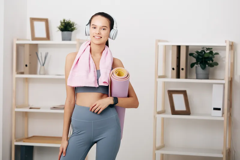 Woman fitness enthusiast in a bright studio, wearing athletic leggings and headphones, with a towel over her shoulder and a yoga mat under arm, ready for a home workout.