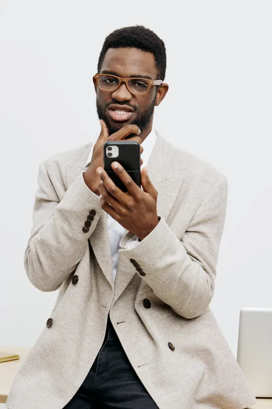 Confident young African-American man in beige blazer and glasses looks surprised while holding a smartphone, isolated on plain white background, studio portrait. Business lifestyle concept